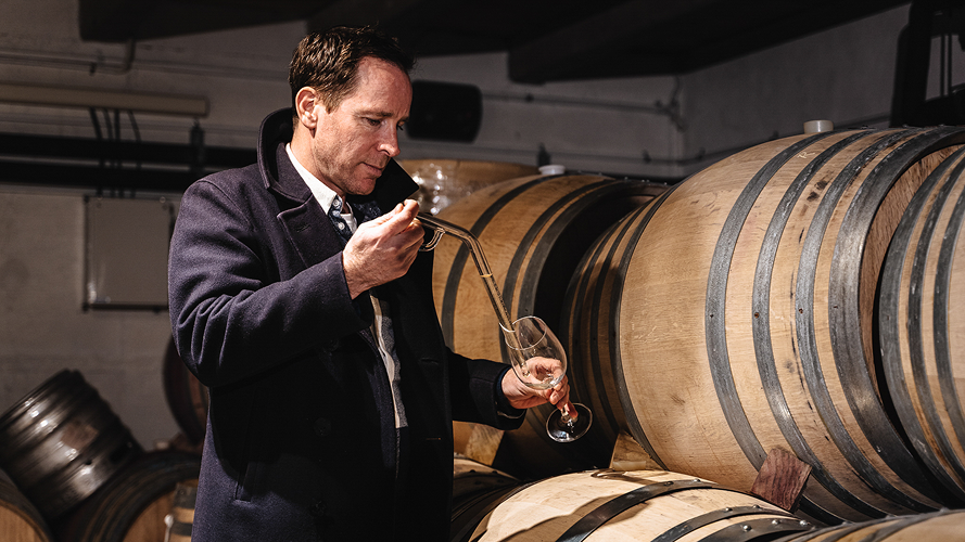 Winemaker Sam Harrop by oak barrels using wine thief